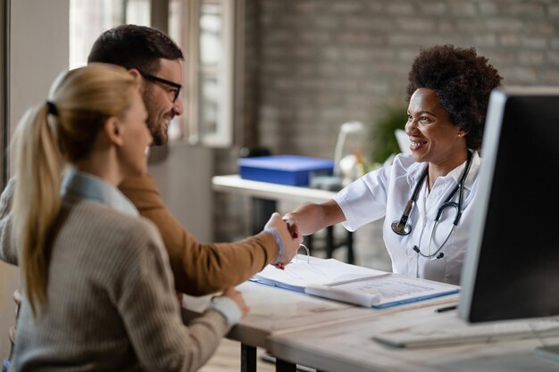 Una pareja feliz dándose la mano con una doctora negra después de las consultas en la clínica El foco está en el doctor