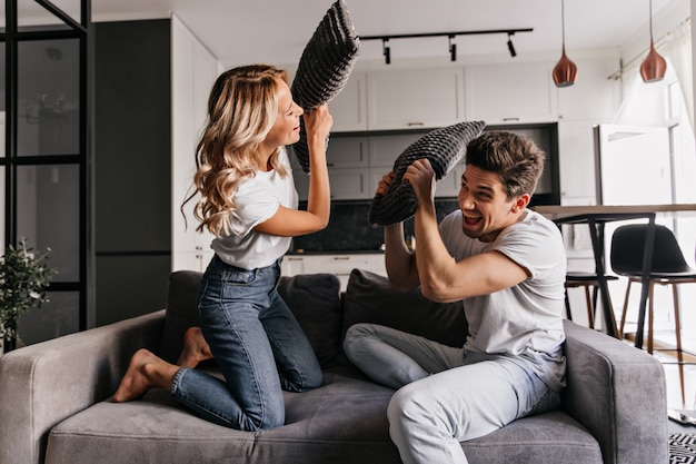 Foto gratuita pareja feliz disfrutando de la pelea de almohadas. chica alegre jugando con su novio en la sala de estar.