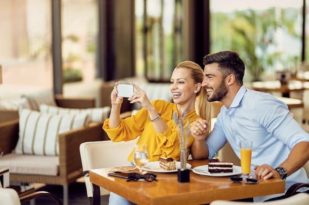 Una pareja feliz divirtiéndose mientras usa un teléfono inteligente y come pastel en un café