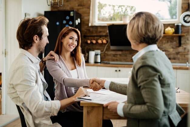Una pareja feliz haciendo un trato con un asesor financiero en una reunión en casa. El foco está en la mujer que le da la mano al asesor.