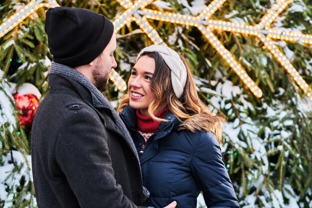 Una pareja feliz con ropa abrigada se toma de la mano y se mira, parada cerca de un árbol de Navidad de la ciudad, disfrutando pasar tiempo juntos.