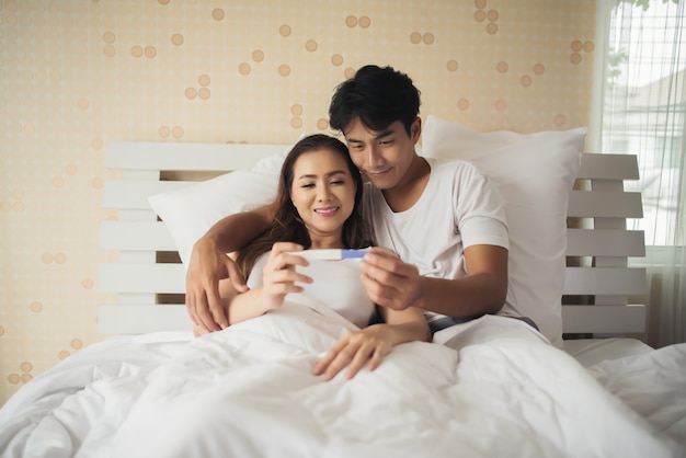 Pareja feliz sonriendo después de descubrir prueba de embarazo positiva en el dormitorio