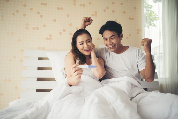 Pareja feliz sonriendo después de descubrir prueba de embarazo positiva en el dormitorio