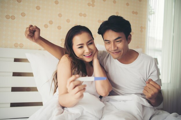 Pareja feliz sonriendo después de descubrir prueba de embarazo positiva en el dormitorio