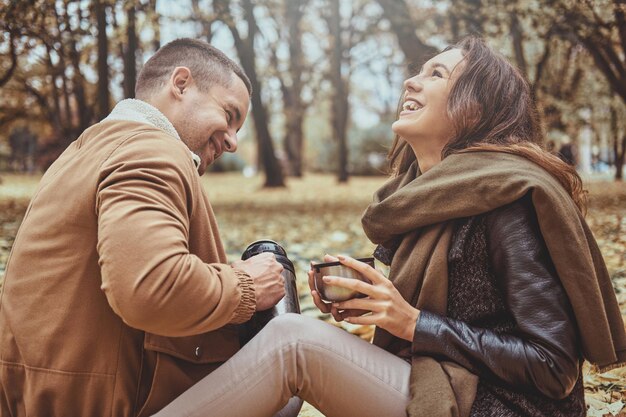 Una pareja feliz sonriente está bebiendo una bebida caliente de un termo mientras se sienta en el parque de otoño.