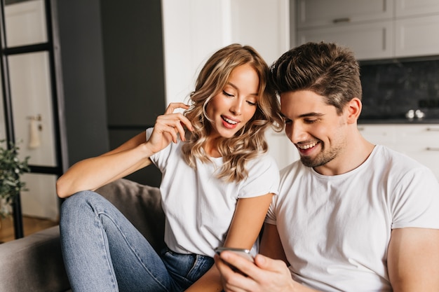 Pareja feliz con teléfono inteligente mirando a la pantalla y sonriendo. Retrato casero de dos jóvenes disfrutando del día juntos y leyendo buenas noticias.