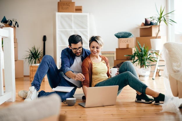 Una pareja feliz usando una laptop mientras se relaja en el piso de su nuevo hogar