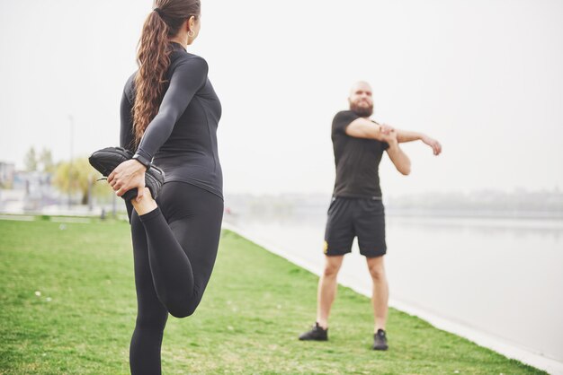 Pareja de fitness estiramientos al aire libre en el parque cerca del agua. Joven barbudo hombre y mujer haciendo ejercicio juntos en la mañana