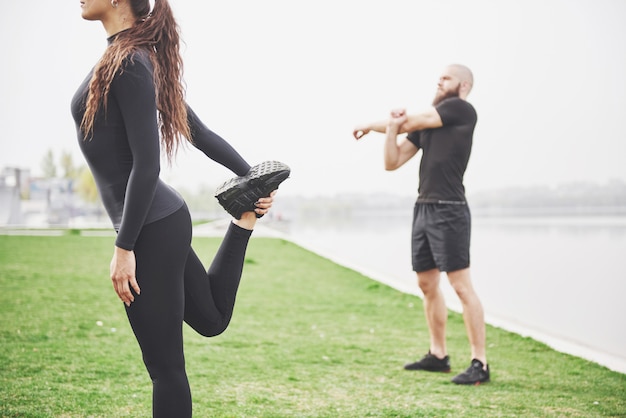 Pareja de fitness estiramientos al aire libre en el parque cerca del agua. joven barbudo hombre y mujer haciendo ejercicio juntos en la mañana