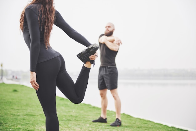 Foto gratuita pareja de fitness estiramientos al aire libre en el parque cerca del agua. joven barbudo hombre y mujer haciendo ejercicio juntos en la mañana