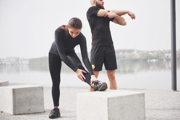 Foto gratuita pareja de fitness estiramientos al aire libre en el parque cerca del agua. joven barbudo hombre y mujer haciendo ejercicio juntos en la mañana