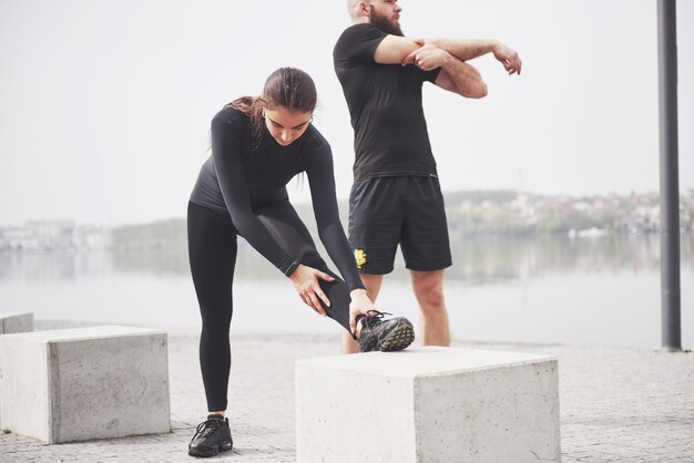 Pareja de fitness estiramientos al aire libre en el parque cerca del agua. Joven barbudo hombre y mujer haciendo ejercicio juntos en la mañana