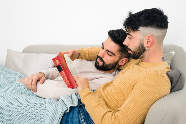 Una pareja gay acostada en el sofá leyendo un libro en casa