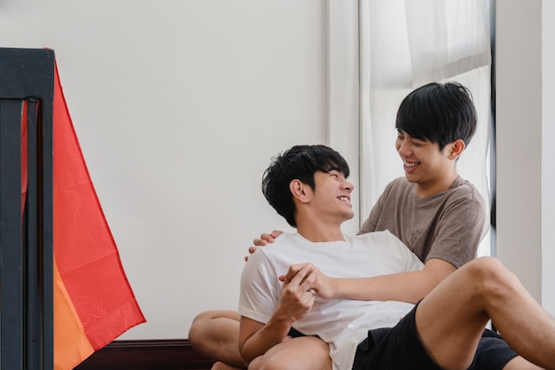 Pareja gay asiática acostado y abrazándose en el piso en casa. Jóvenes asiáticos LGBTQ + hombres besándose felices relajarse descansar juntos pasar tiempo romántico en la sala de estar con la bandera del arco iris en la casa moderna de la mañana.