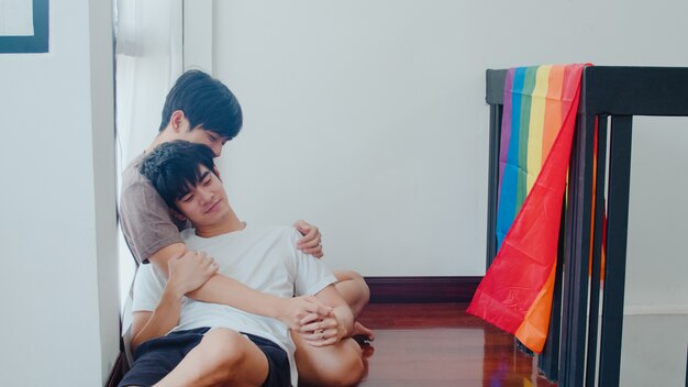 Pareja gay asiática acostado y abrazándose en el piso en casa. Jóvenes asiáticos LGBTQ + hombres besándose felices relajarse descansar juntos pasar tiempo romántico en la sala de estar con la bandera del arco iris en la casa moderna de la mañana.