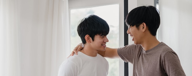 Pareja gay asiática de pie y abrazando cerca de la ventana en casa. Jóvenes asiáticos LGBTQ + hombres besándose felices relajarse descansar juntos pasar tiempo romántico en la sala de estar en casa moderna en la mañana.