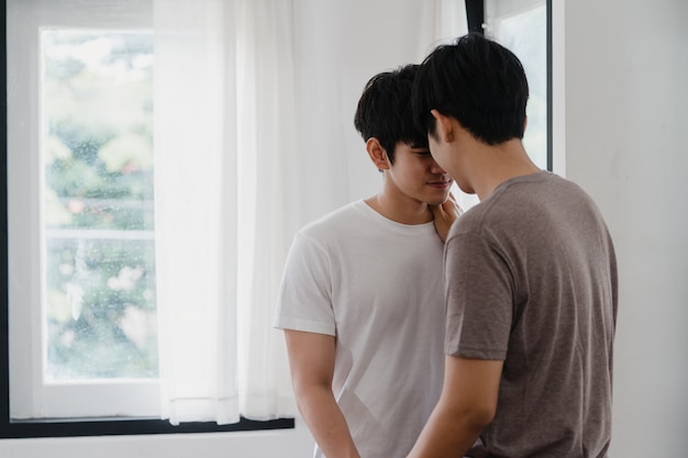 Pareja gay asiática de pie y abrazando cerca de la ventana en casa. Jóvenes asiáticos LGBTQ + hombres besándose felices relajarse descansar juntos pasar tiempo romántico en la sala de estar en casa moderna en la mañana.
