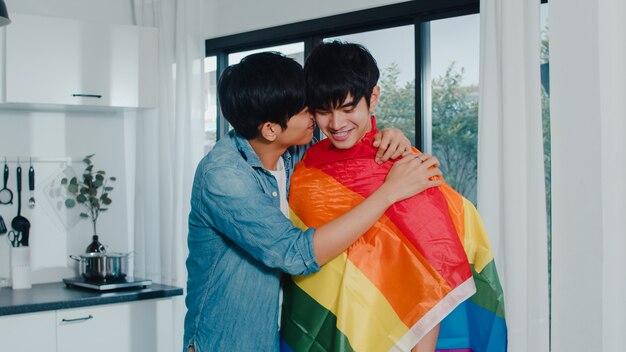 Pareja gay asiática de pie y abrazos habitación en casa. Los jóvenes y guapos hombres LGBTQ + besándose felices se relajan descansan juntos pasan tiempo romántico en la cocina moderna con la bandera del arco iris en la casa por la mañana.