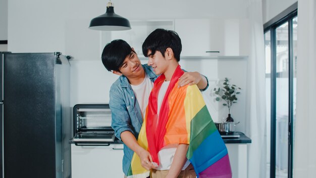 Pareja gay asiática de pie y abrazos habitación en casa. Los jóvenes y guapos hombres LGBTQ + besándose felices se relajan descansan juntos pasan tiempo romántico en la cocina moderna con la bandera del arco iris en la casa por la mañana.