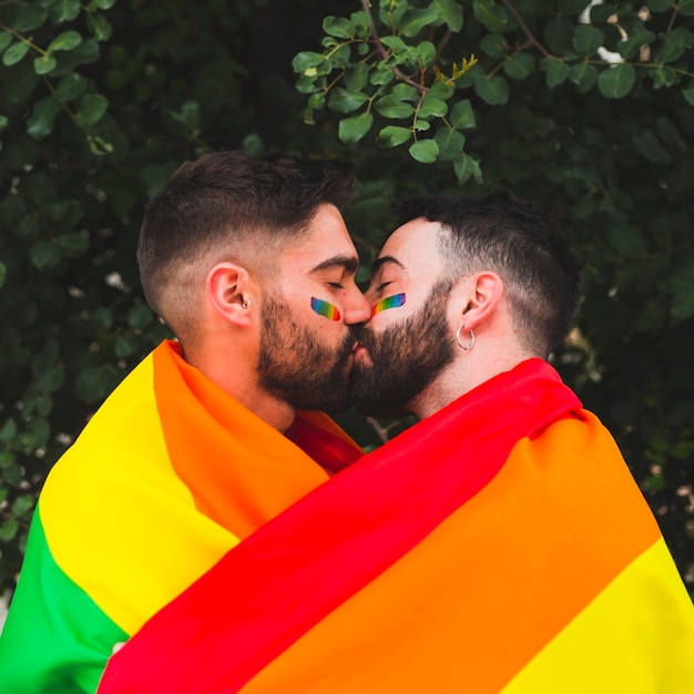 Pareja gay besándose con la bandera del arco iris en el parque