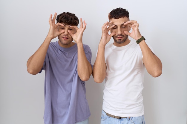 Una pareja gay homosexual de pie sobre un fondo blanco tratando de abrir los ojos con los dedos, soñolienta y cansada por la fatiga matutina