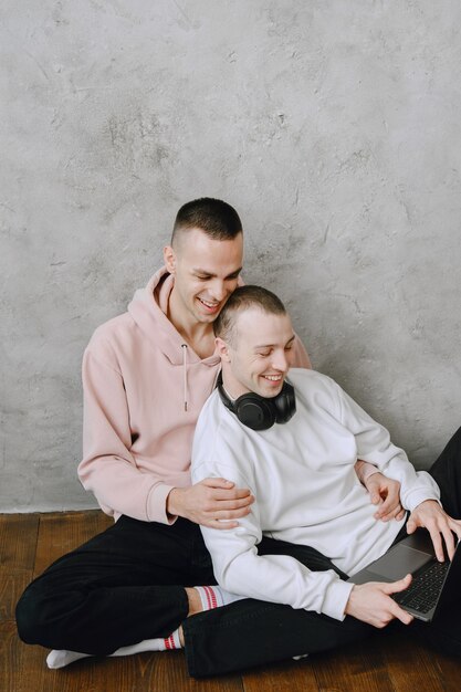 Una pareja gay joven sentada en el piso usando una computadora portátil, usando auriculares, escuchan música juntos, abrazándose o abrazándose.