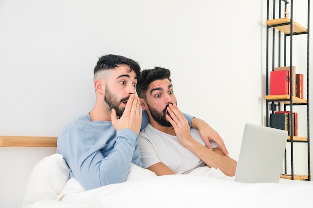 Foto gratuita pareja gay joven sorprendida que se sienta en la cama que mira el ordenador portátil