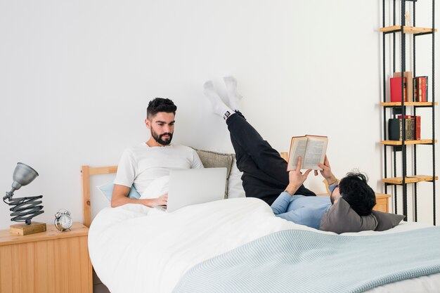 Pareja gay joven tumbada en la cama leyendo el libro y usando el teléfono móvil