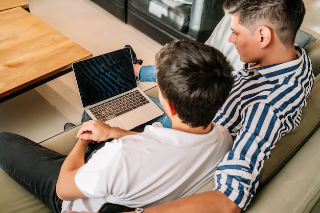 Una pareja gay pasando tiempo juntos y usando una computadora portátil mientras está sentado en un sofá en casa.