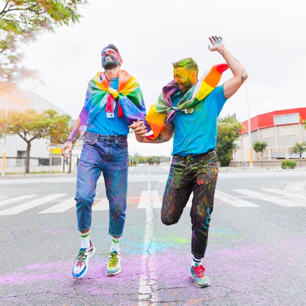 Pareja gay riendo corriendo en la carretera con la bandera del arco iris