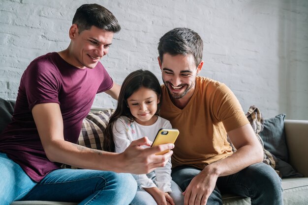 Una pareja gay y su hija usando un teléfono móvil en casa.