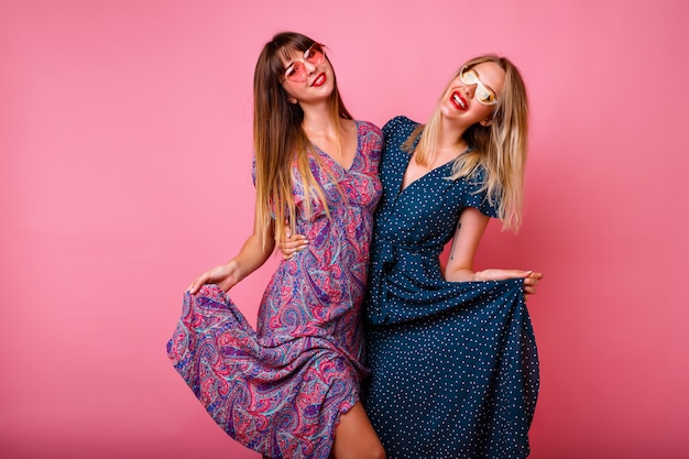 Pareja de hermanas de amigas bonitas divirtiéndose juntas, vistiendo elegantes vestidos y gafas de sol de moda de verano, posando en la pared rosa, abrazos y sonriendo, ambiente de fiesta.