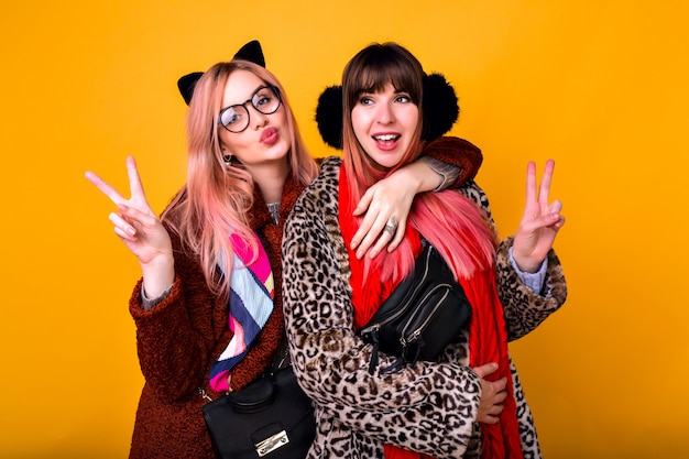 Pareja de hipster bastante divertidas mejores amigas hermanas chicas haciendo selfie en la pared amarilla, mostrando la lengua y sonriendo, vistiendo abrigos de piel estampados de primavera, bufandas, riñonera y gafas transparentes.