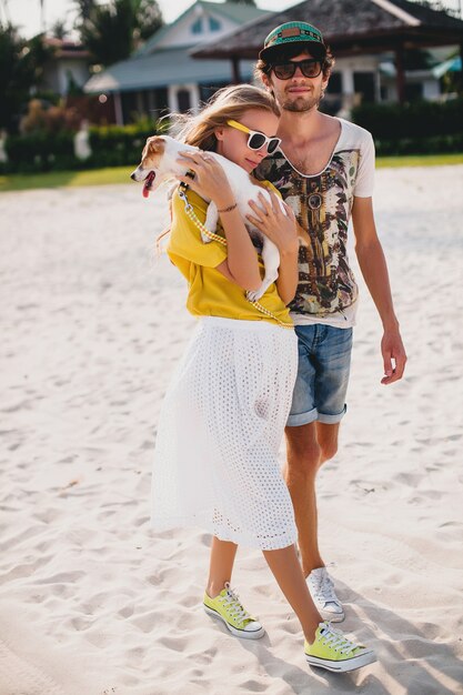 Pareja de hipster con estilo joven inconformista en el amor caminando y jugando con el perro en la playa tropical