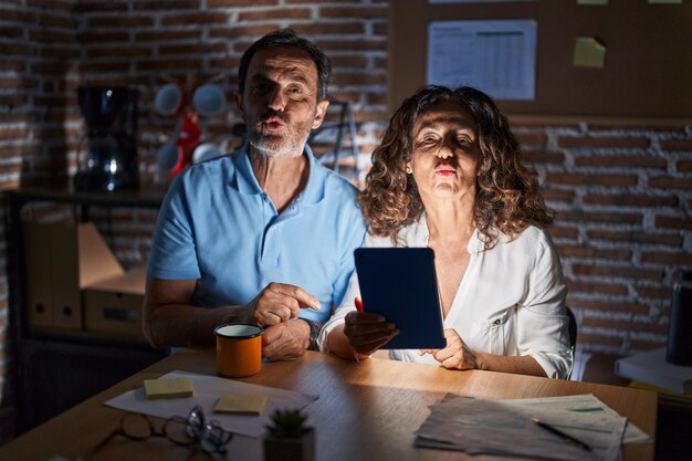 Una pareja hispana de mediana edad usando un panel táctil sentado en la mesa por la noche mirando a la cámara lanzando un beso al aire siendo encantadora y sexy. expresión de amor.