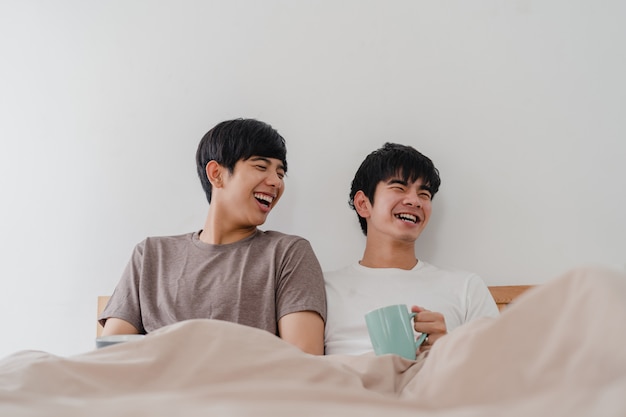 Pareja de hombres gays asiáticos hablando pasar un buen rato en el hogar moderno. Joven Asia amante masculino feliz relajarse descansar beber café después de despertarse mientras está acostado en la cama en el dormitorio en la casa por la mañana.