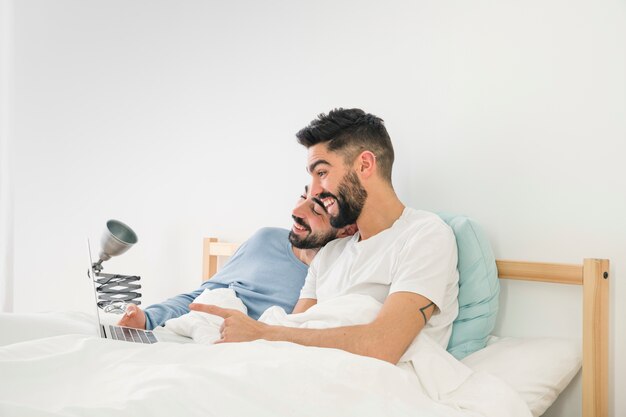 Pareja de homosexuales acostada en la cama riéndose mientras mira un portátil contra una pared blanca