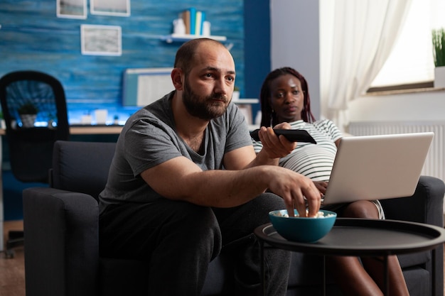 Pareja interracial embarazada esperando bebé en casa. Mujer afroamericana con portátil moderno mientras hombre caucásico sosteniendo el control remoto de la TV y comiendo palomitas de maíz. Familia de raza mixta