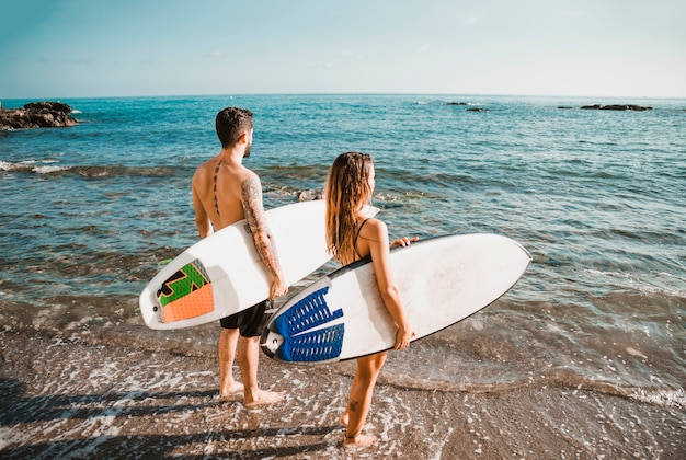 Pareja irreconocible con tablas de surf de pie cerca del mar agitando