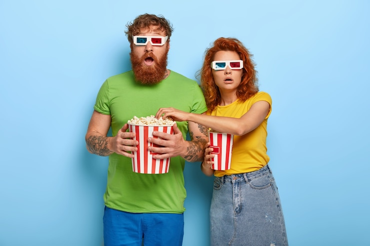 Foto gratuito pareja de jengibre asustada ve una película de miedo, mira con estupefacción, come palomitas de maíz, usa anteojos estéreo