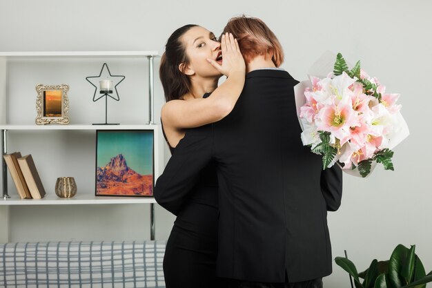 Pareja joven se abrazó el día de la mujer feliz con ramo de flores susurros de mujer de pie en la sala de estar