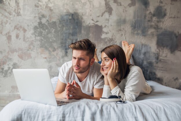 Pareja joven acostado en la cama en casa en ropa casual, mirando en la computadora portátil, pasar tiempo juntos, mujer romántica, hombre serio