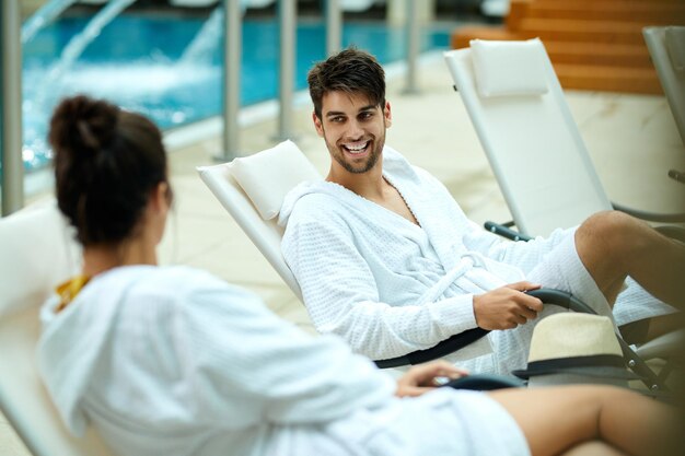 Pareja joven en albornoces hablando mientras está sentado junto a la piscina y pasando un fin de semana en el spa El foco está en el hombre feliz