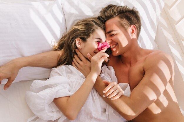 Pareja joven alegre en el amor acostado en la cama blanca, disfrutando de la luna de miel en la mañana soleada. Hombre guapo sonriente con piel bronceada abrazando a su magnífica novia en blusa vintage descansando sobre almohadas