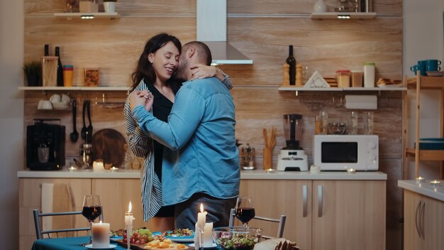 Pareja joven alegre bailando y besándose durante una cena romántica. Feliz pareja de enamorados cenando juntos en casa, disfrutando de la comida, celebrando su aniversario.