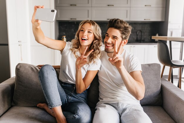 Pareja joven alegre en casa haciendo selfie con signo de la paz y sonriendo ampliamente. Niña feliz sentada en el sofá con su novio y tomando un retrato de ellos juntos.