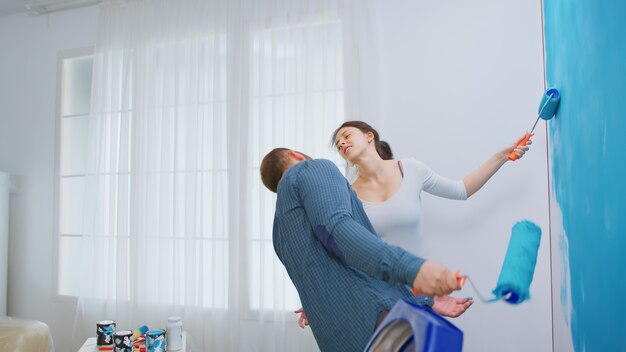 Pareja joven alegre que decora el apartamento y el baile. Divertirse y pintar las paredes. Redecoración de apartamentos y construcción de viviendas mientras se renueva y mejora. Reparación y decoración.