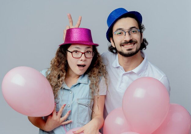 Foto gratuita pareja joven alegre con sombrero rosa y azul chico haciendo orejas de conejo gesto a chica sosteniendo globos aislado sobre fondo blanco.