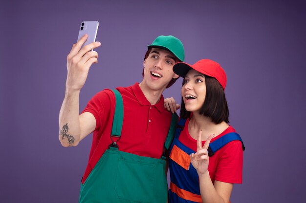 Pareja joven alegre en uniforme de trabajador de la construcción y gorra tomando selfie juntos chica poniendo la mano sobre el hombro del chico haciendo el signo de la paz