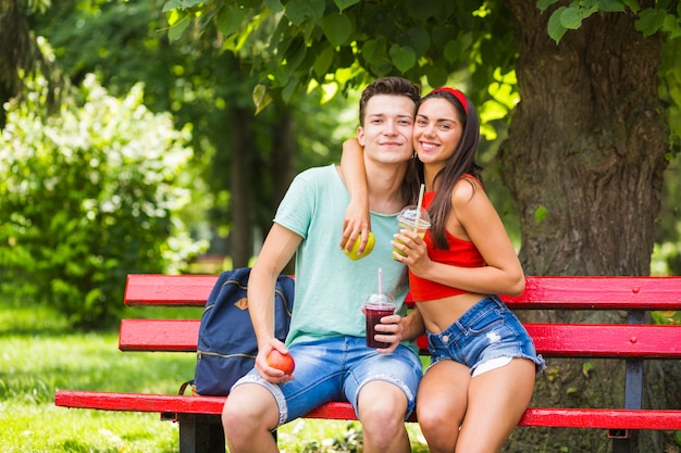 Pareja joven amistosa con batidos y manzanas sentado en el banco